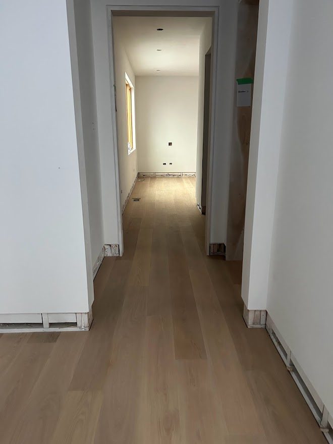 A hallway into a white unfinished bedroom where the baseboards are missing and the hardwood flooring is brand new.