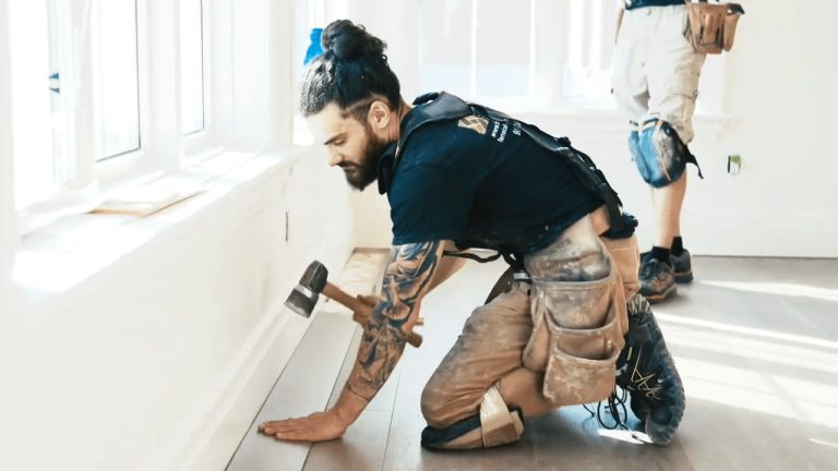 A carpenter kneeling down in knee pads using his hammer to place hardwood flooring into the bright white room.